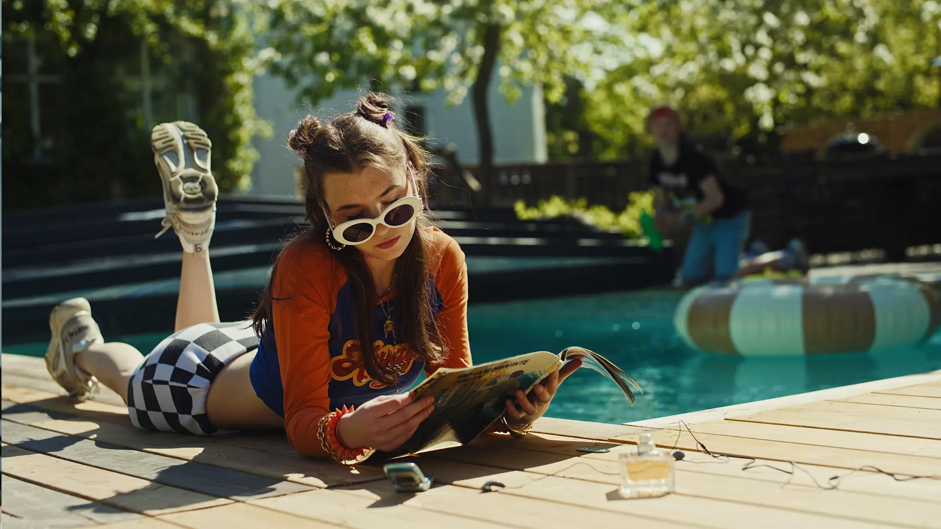 En ung flicka med solglasögon läser en tidning vid poolkanten